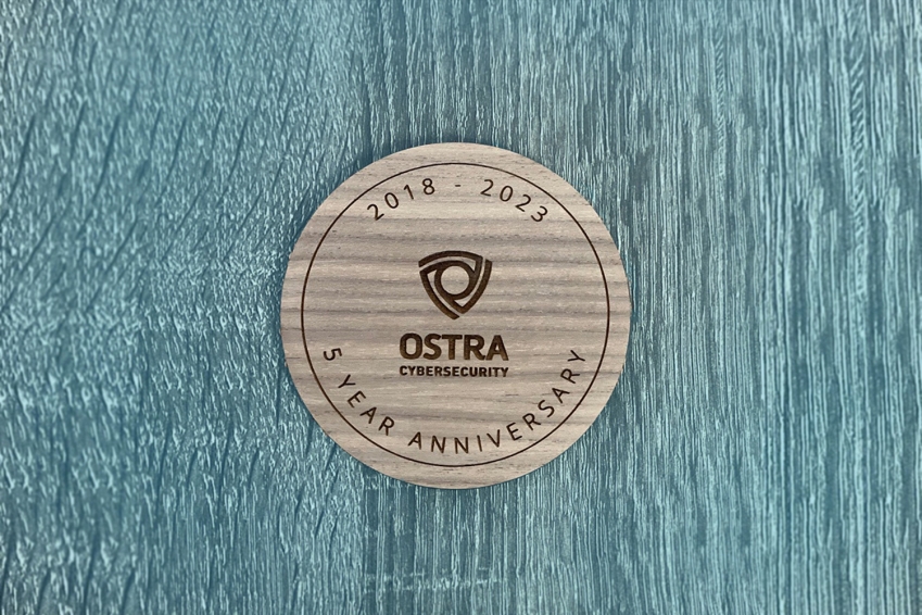 round wood plaque on blue stained wood background commemorating Ostra Cybersecurity's 5 year anniversary